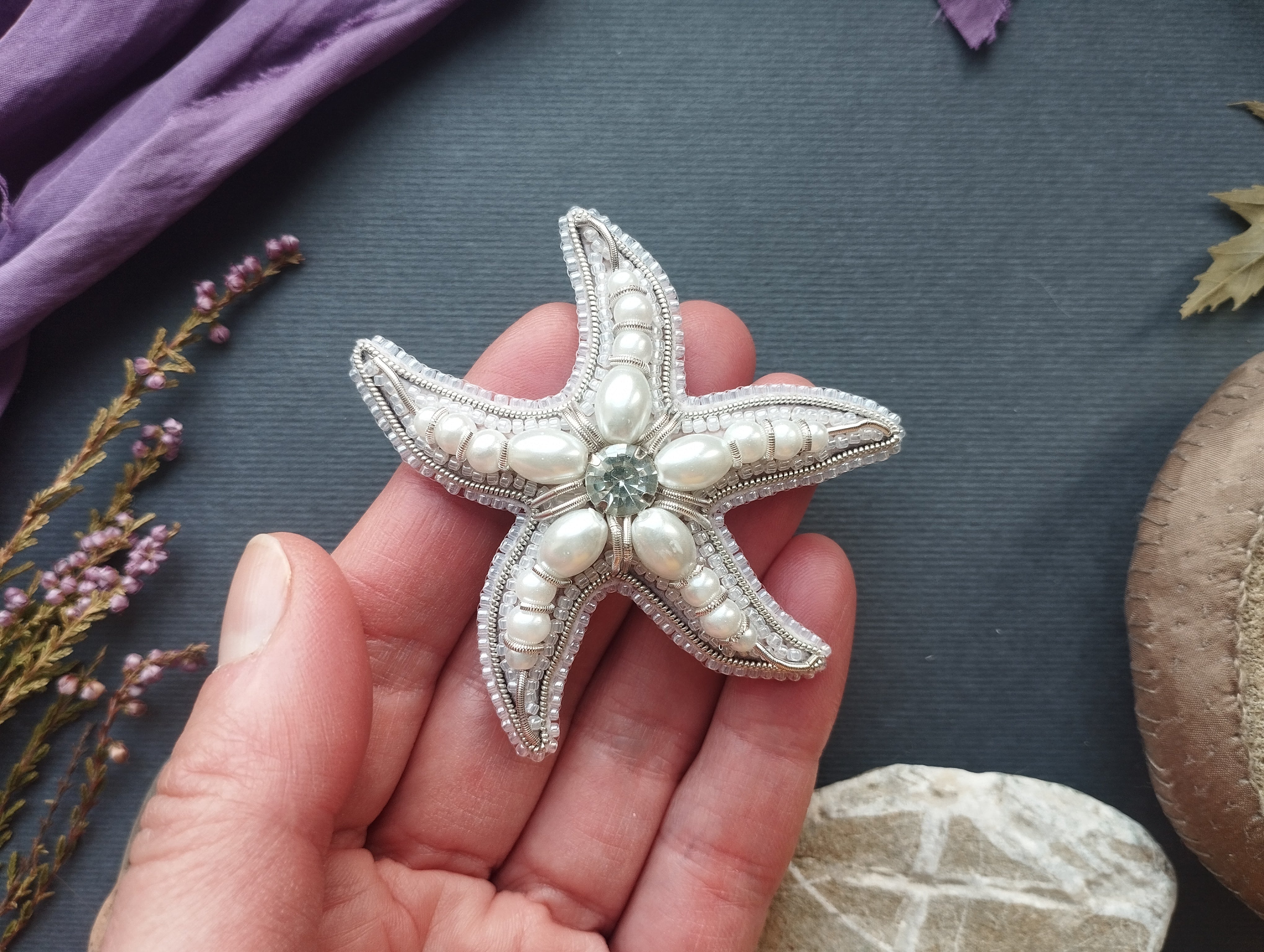 A hand holding a decorative starfish with pearls and a gemstone on a dark background.